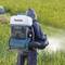 A person wearing a protective suit is operating a portable leaf blower with a white container, surrounded by green plants on a field.