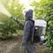 A person is wearing protective clothing and spraying plants in an orchard using a portable device.