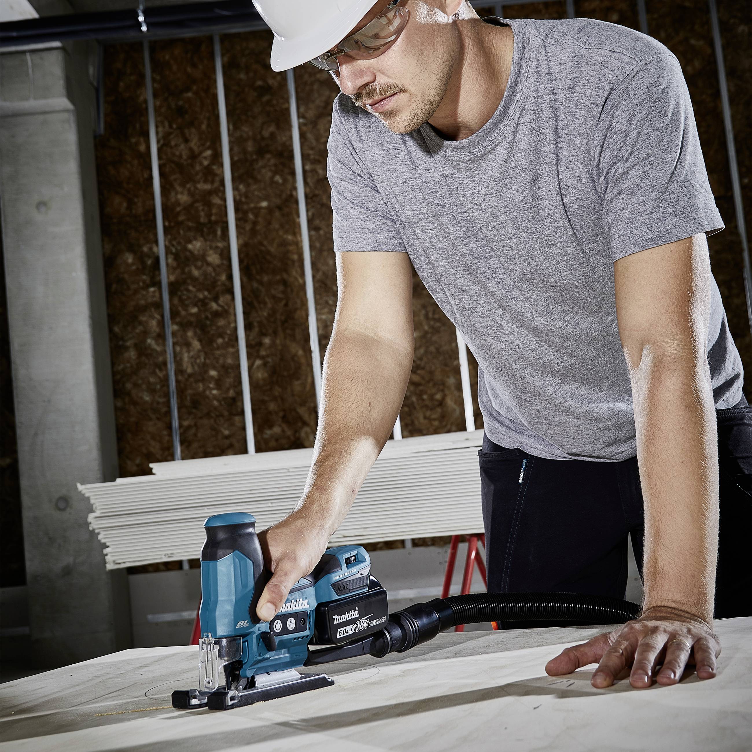 A man wearing safety glasses and a hard hat is working with an electric jigsaw on a wooden board in a workshop.