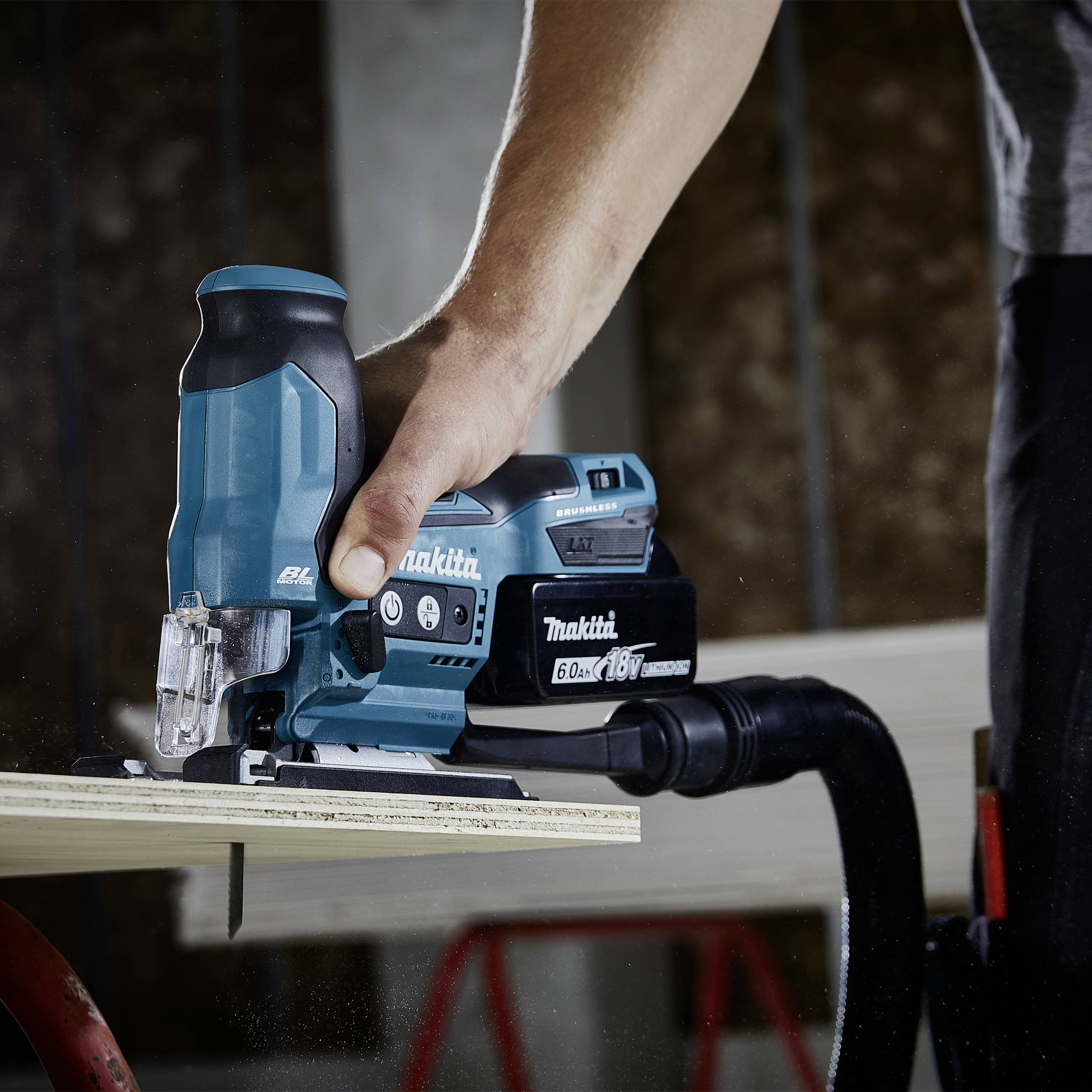 A person is using an electric jigsaw to cut a piece of wood. The jigsaw is blue and connected to a vacuum cleaner.