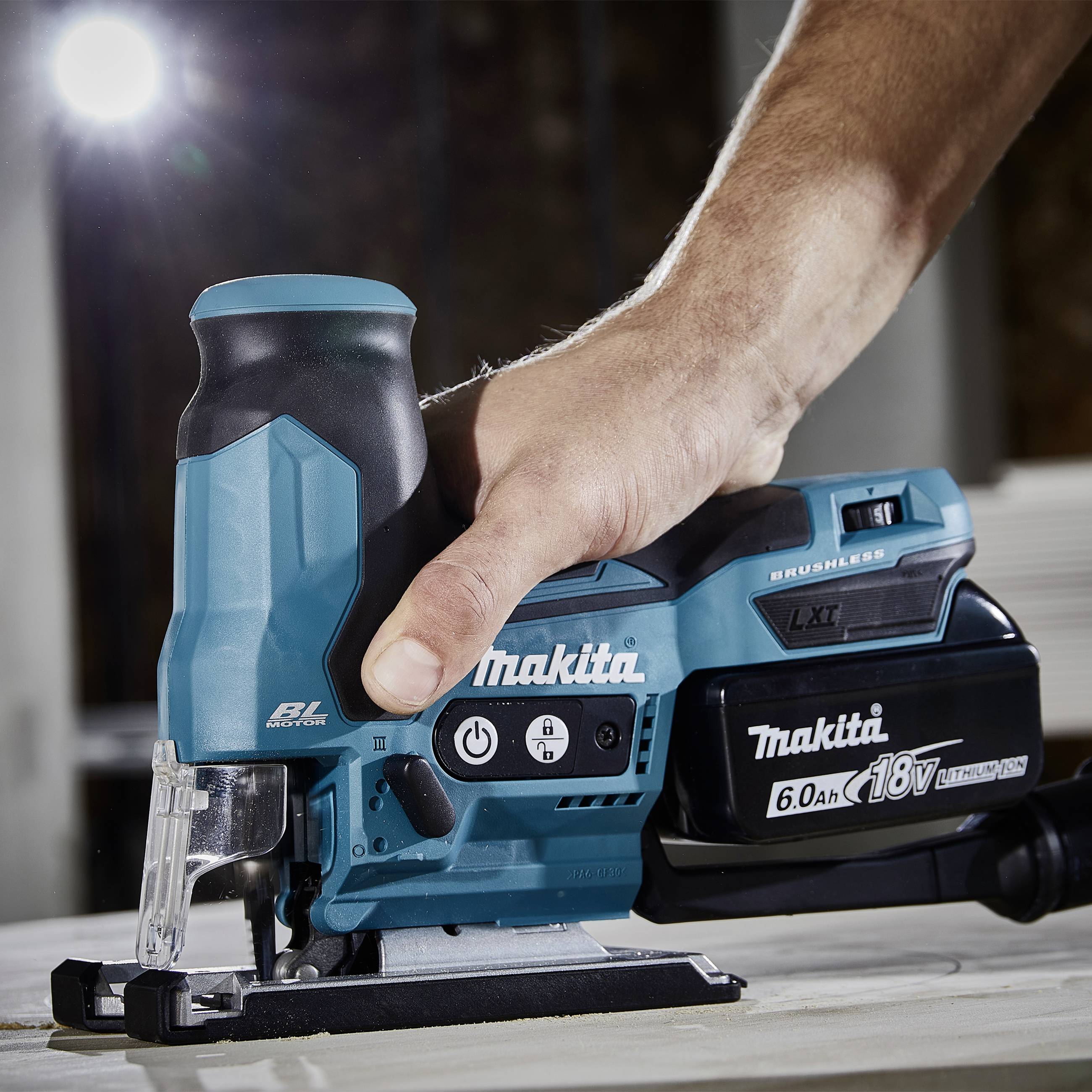 A person is using a blue, battery-powered jigsaw by Makita on a wooden table. They are holding the saw securely with one hand.