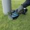 A person is using a portable, battery-powered garden tool to cut grass around a metal post.