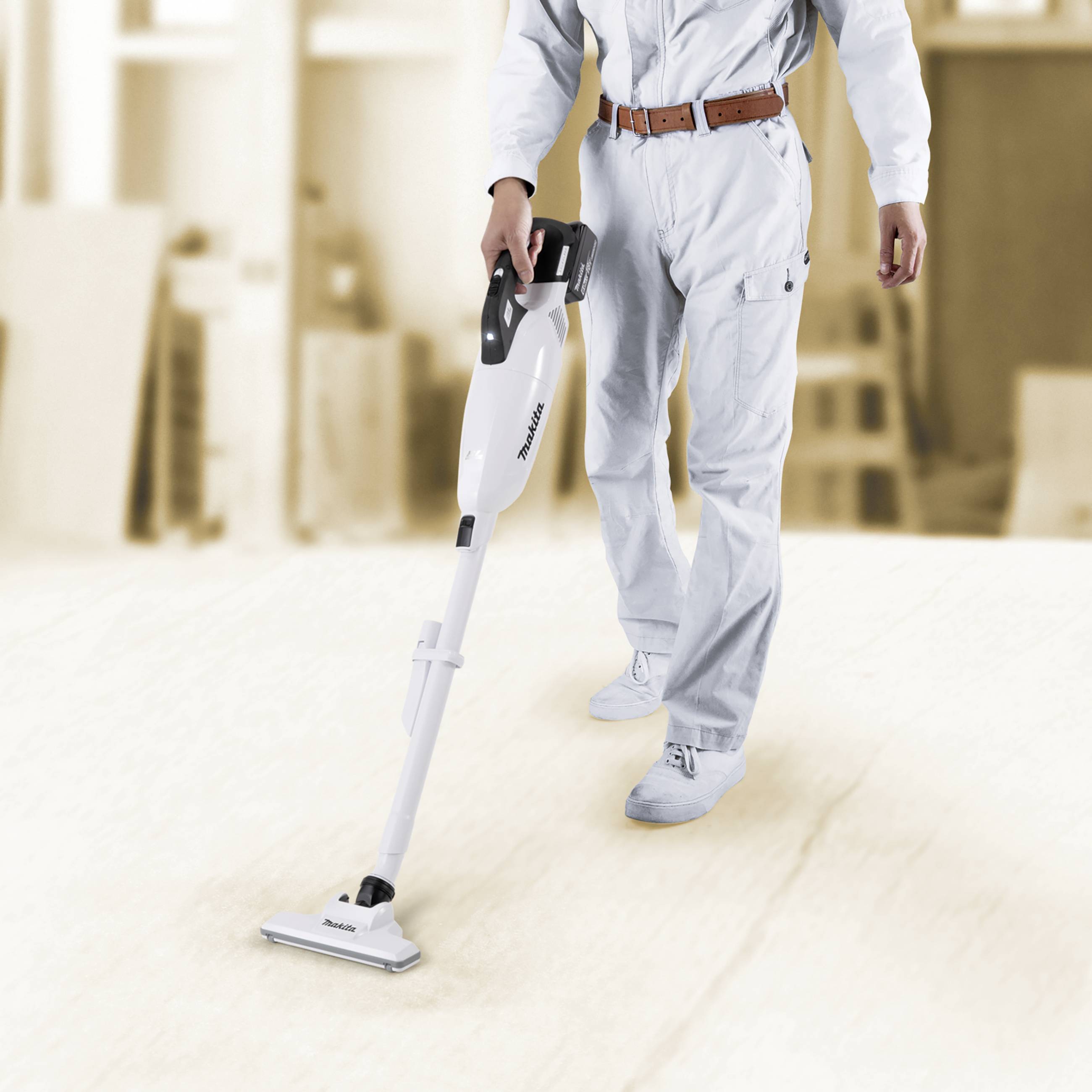 A person in a white overall is vacuuming the floor with a white hoover in a bright room.