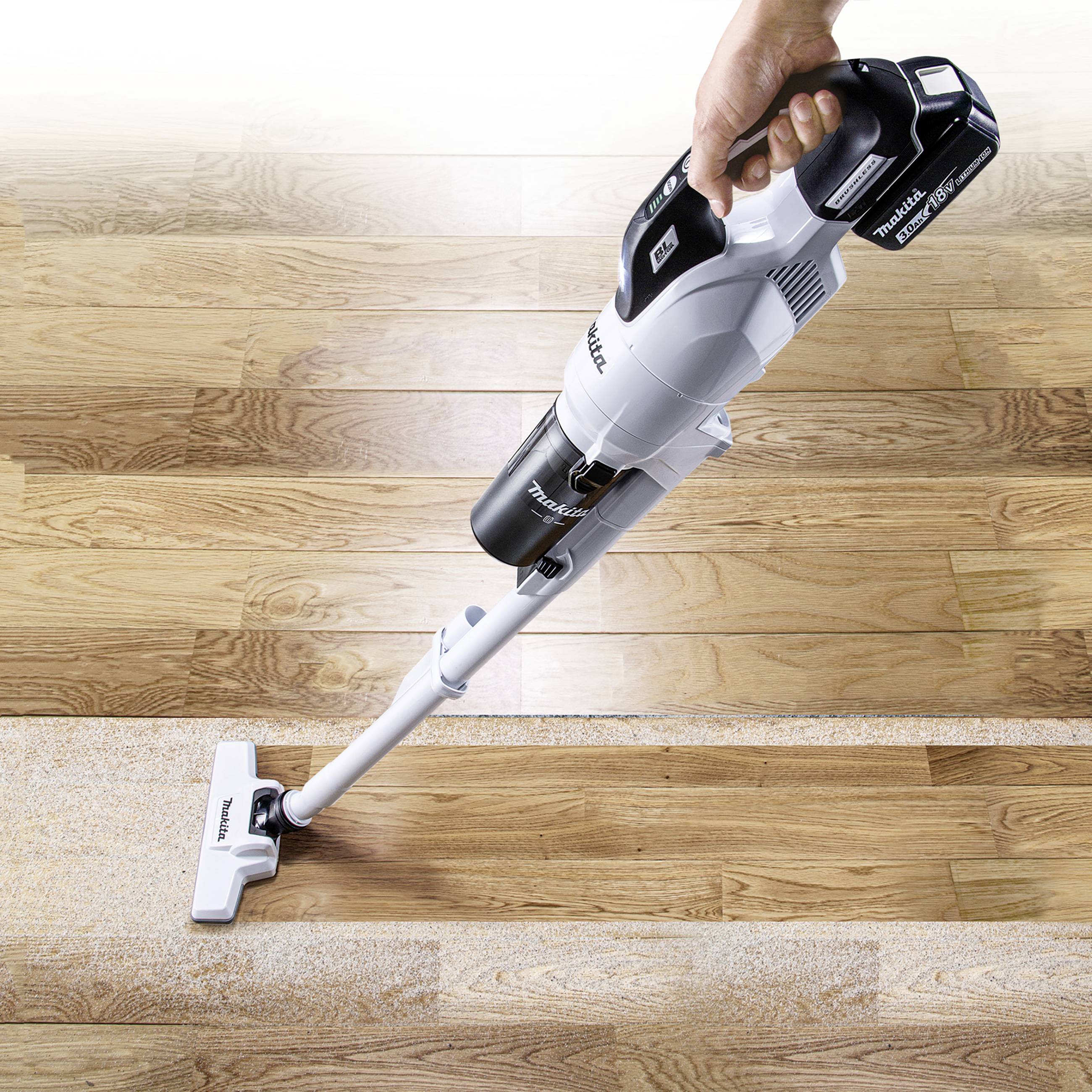 A person is using a cordless vacuum cleaner to remove dirt from a wooden floor, with a clearly visible cleaned area.