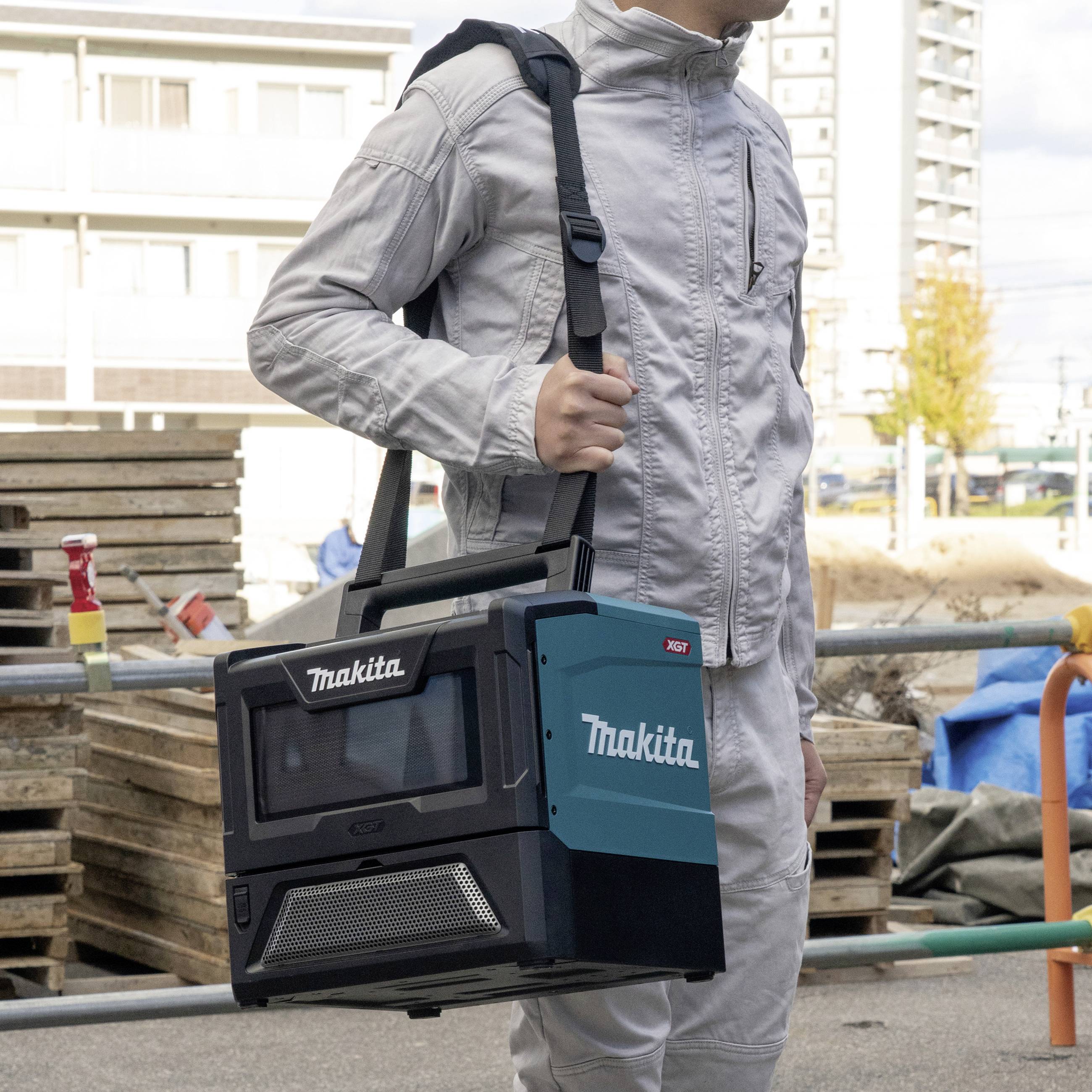 A person is carrying a portable, battery-powered Makita fan heater on a construction site. Construction equipment is visible in the background.