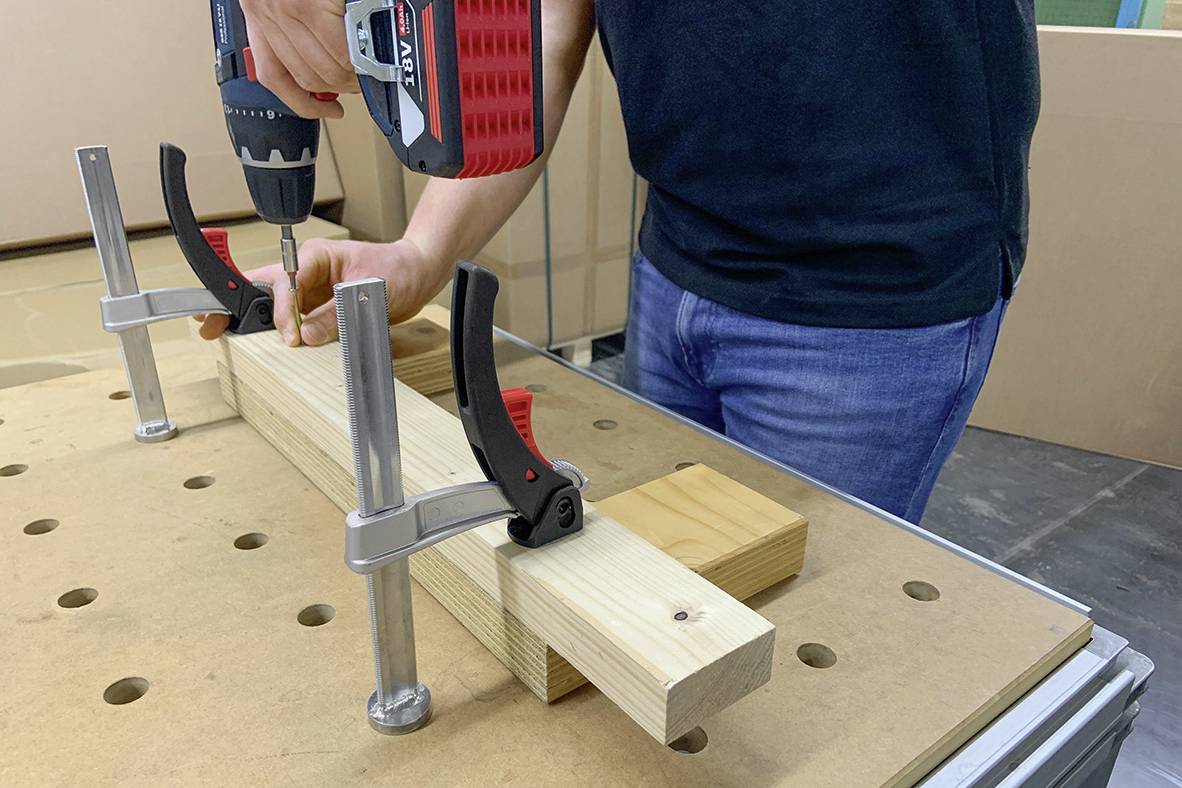 A person secures a piece of wood on a table with clamps and drills a hole.