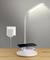 A wireless charger on a table is charging a smartphone under an LED lamp that is plugged into a power socket.