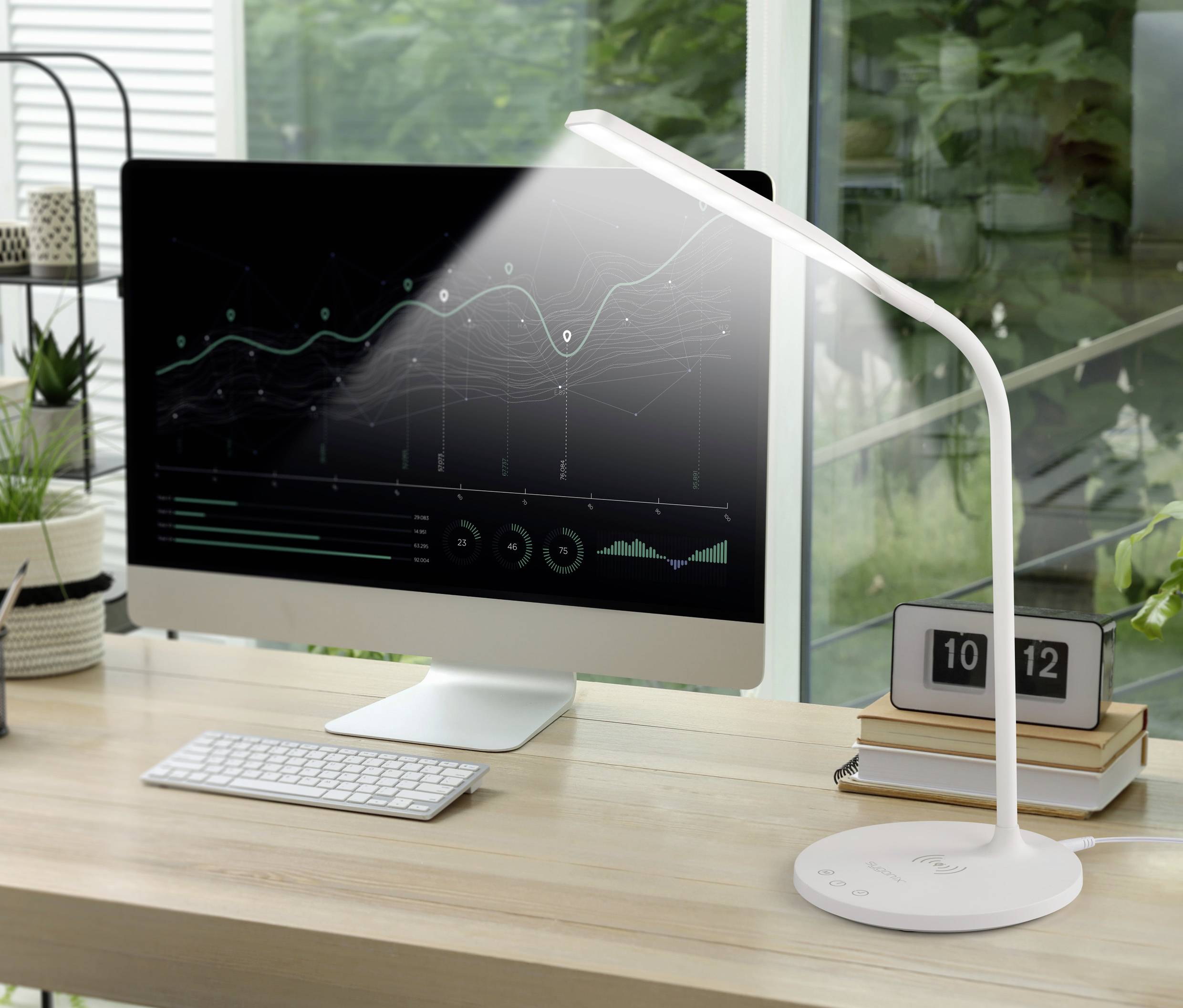 A modern desk with a computer screen displays a diagram with rising curves, next to a lamp and digital clock.