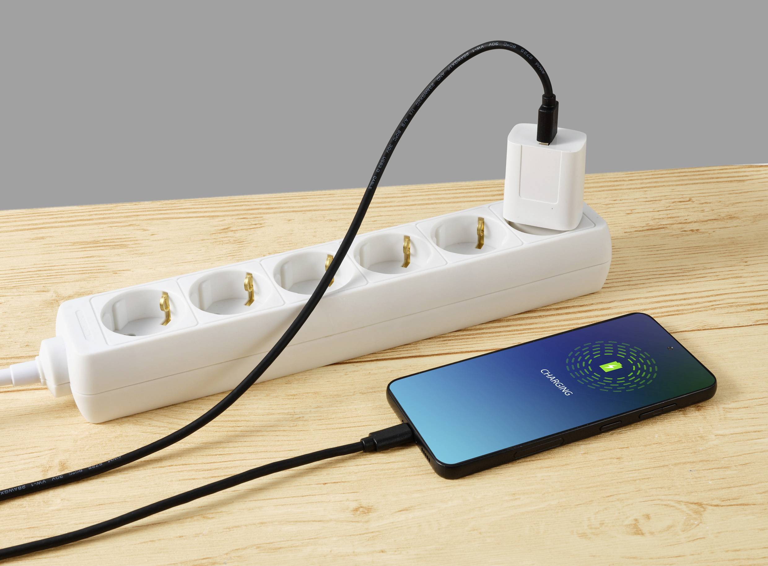 A mobile phone is lying on a table and being charged via a cable connected to a power strip. The display reads 'Charging'.