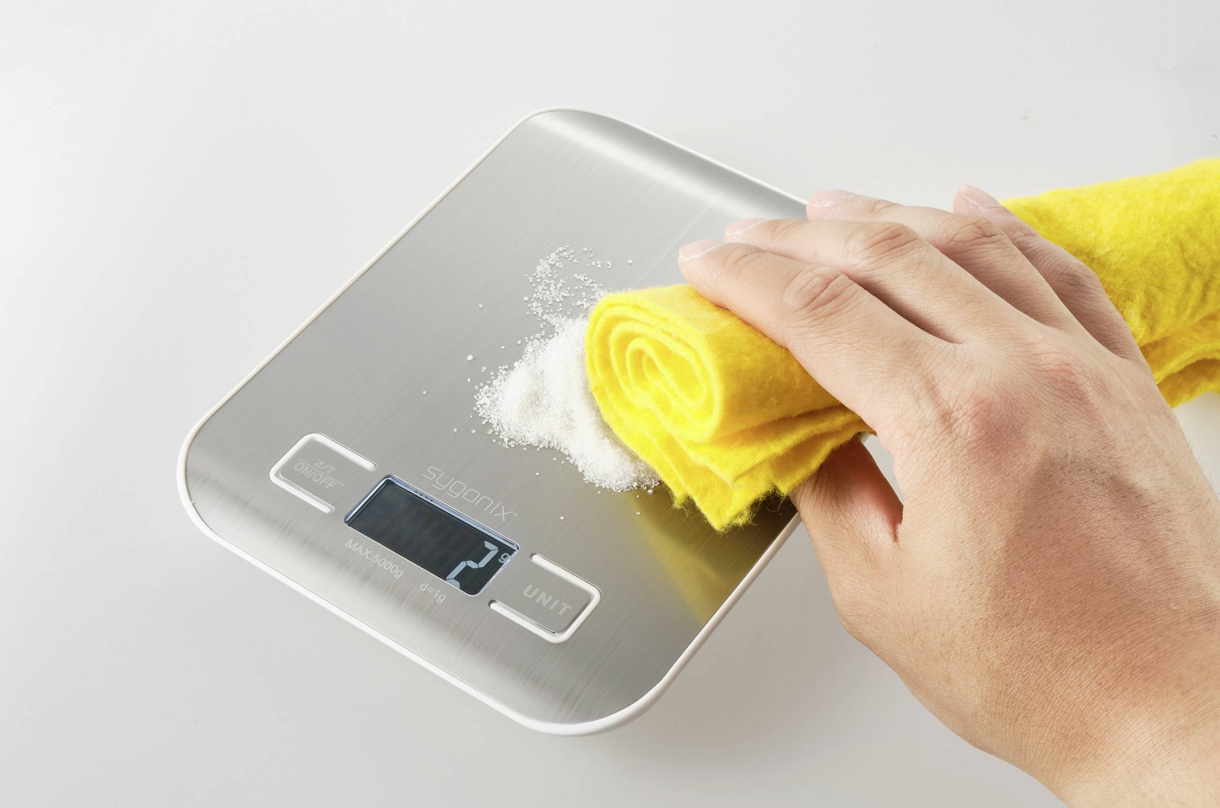 A hand wipes white material from a digital kitchen scale with a yellow cloth; demonstrates the cleaning process and contributes to cleanliness.