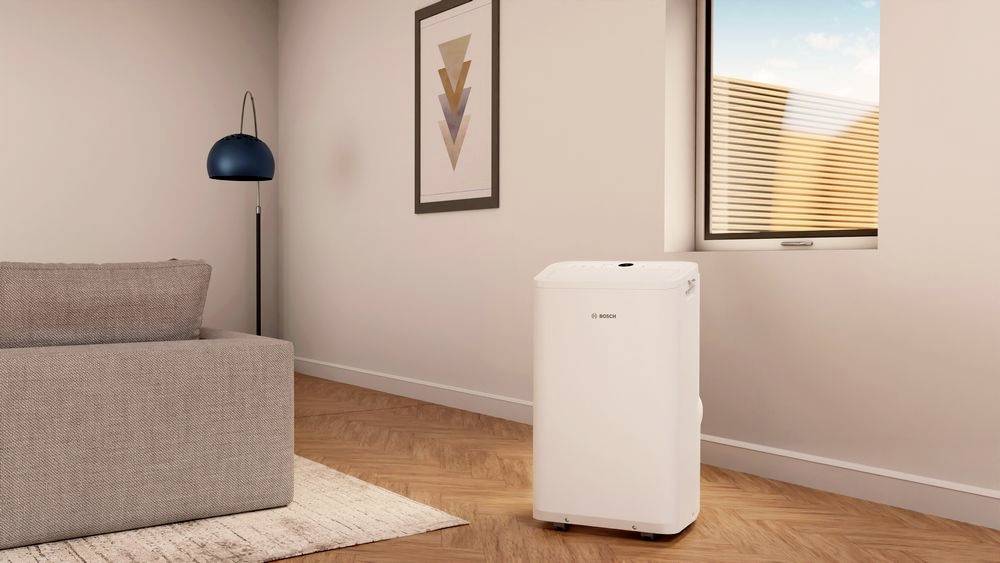 A white dehumidifier stands in a modernly furnished living room with a grey sofa, wooden flooring, and a large window.