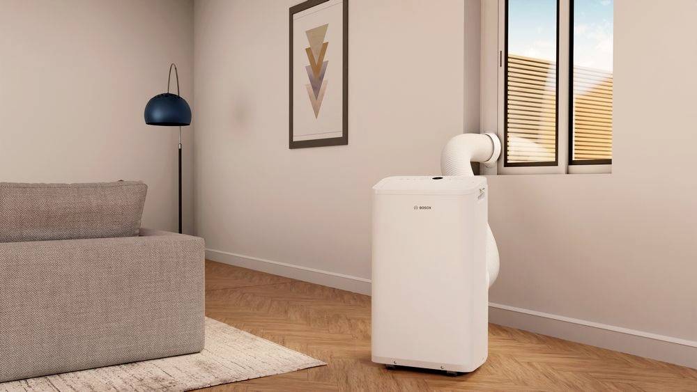 Portable air conditioning unit in a modern living room, connected to a window. A standard lamp stands beside a grey sofa.