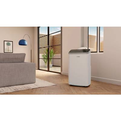 A portable air conditioning unit stands in a modern living room with a wooden floor, next to a grey sofa and large windows overlooking a terrace.
