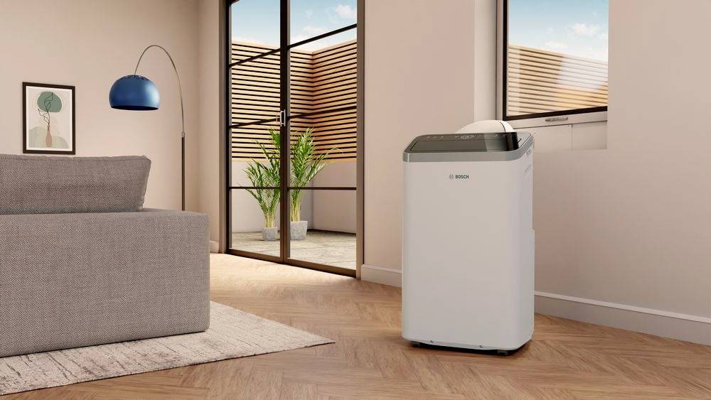 A portable air conditioning unit stands in a modern living room with a wooden floor and large windows. A plant is in the background.