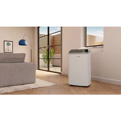 A portable air conditioning unit stands in a modern living room with a wooden floor and large windows. A plant is in the background.