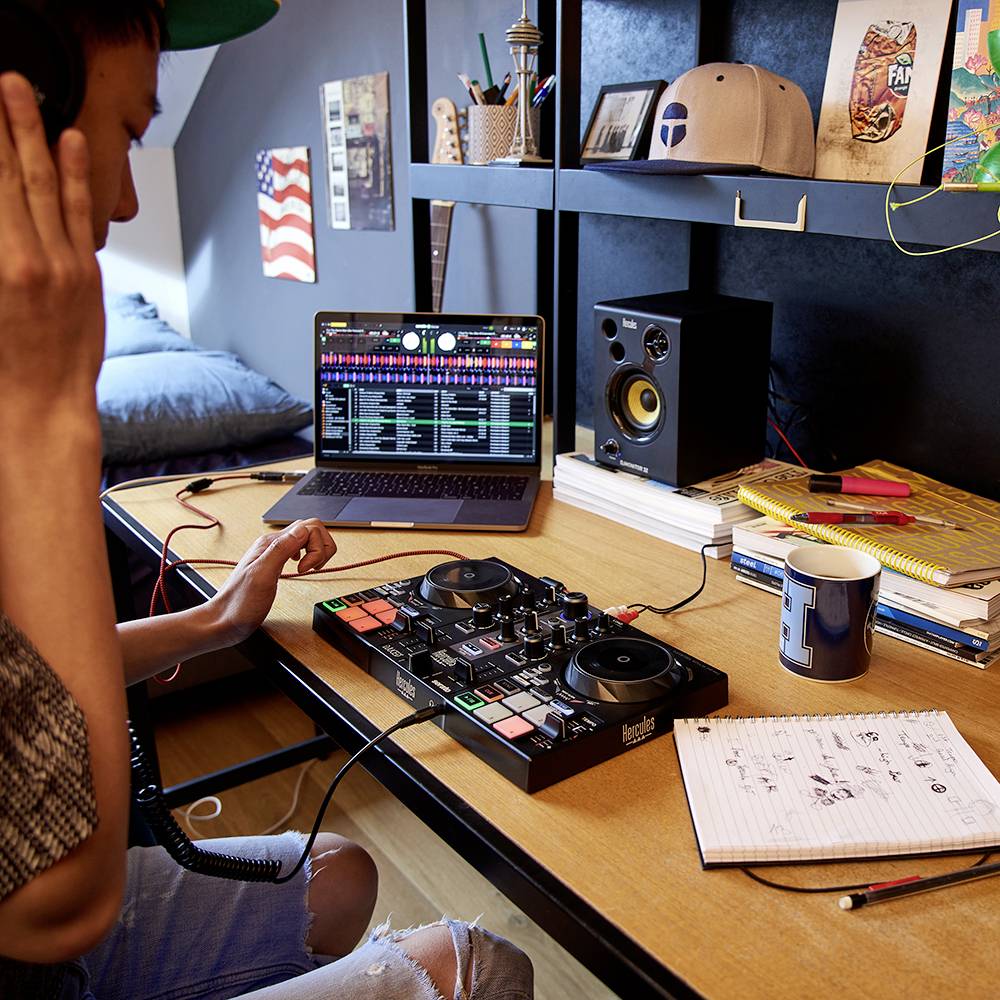 A person is sitting at a table with DJ equipment, headphones and a laptop. A notebook and a mug are visible next to a speaker.