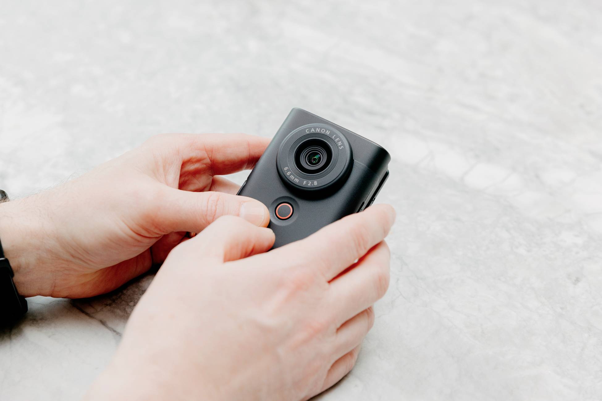 A person is holding a small, black camera in their hands on a grey, marbled surface.