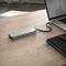 A laptop sits on a wooden table next to a notebook. Connected to it is a USB hub with multiple ports, including USB and HDMI.