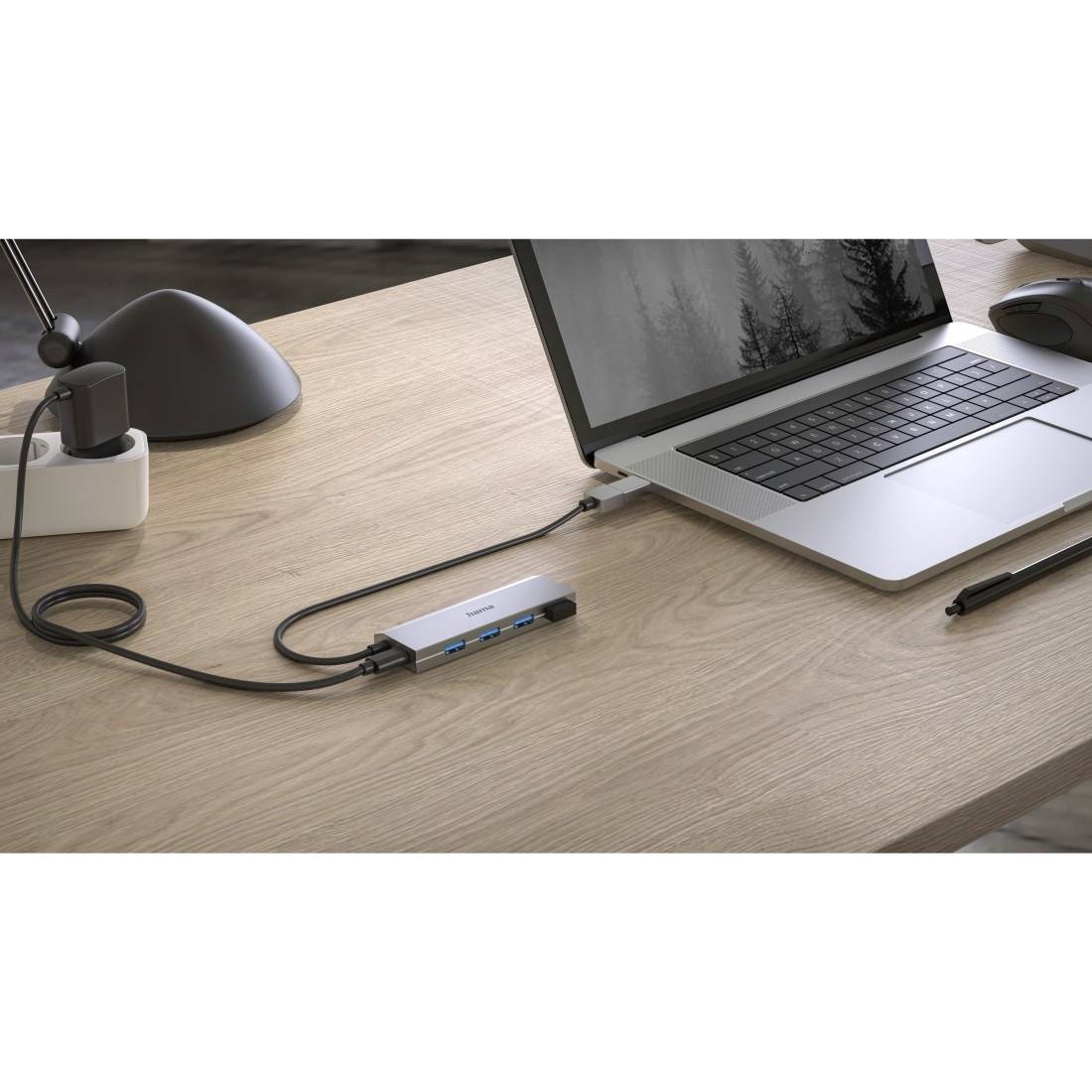 A laptop on a table, connected to a docking station via a cable. Alongside are a pen, lamp, and power socket in the background.