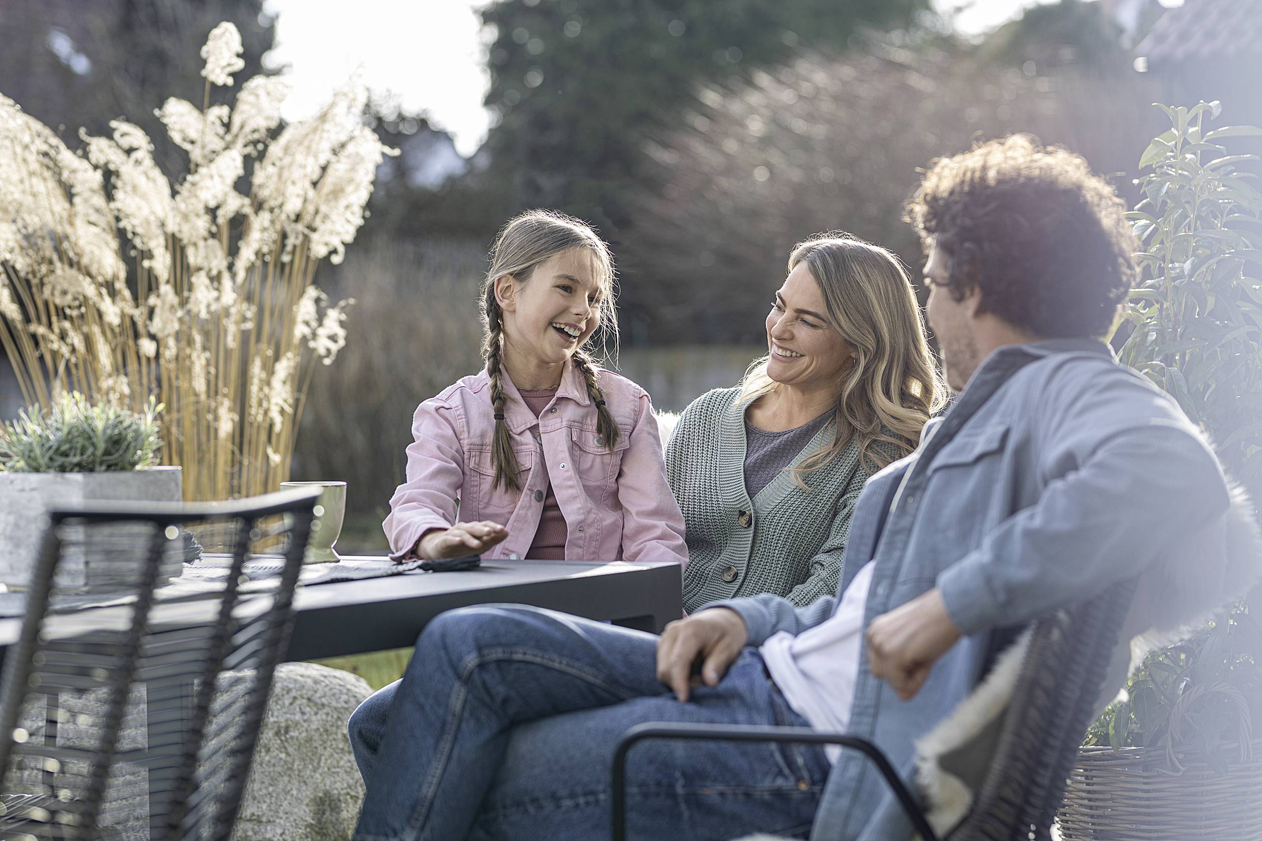 A family is sitting outside at a table. A girl is laughing with a man and a woman. Plants and trees are visible in the background.