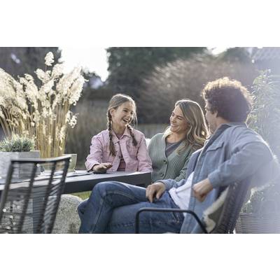 A family is sitting outside at a table. A girl is laughing with a man and a woman. Plants and trees are visible in the background.