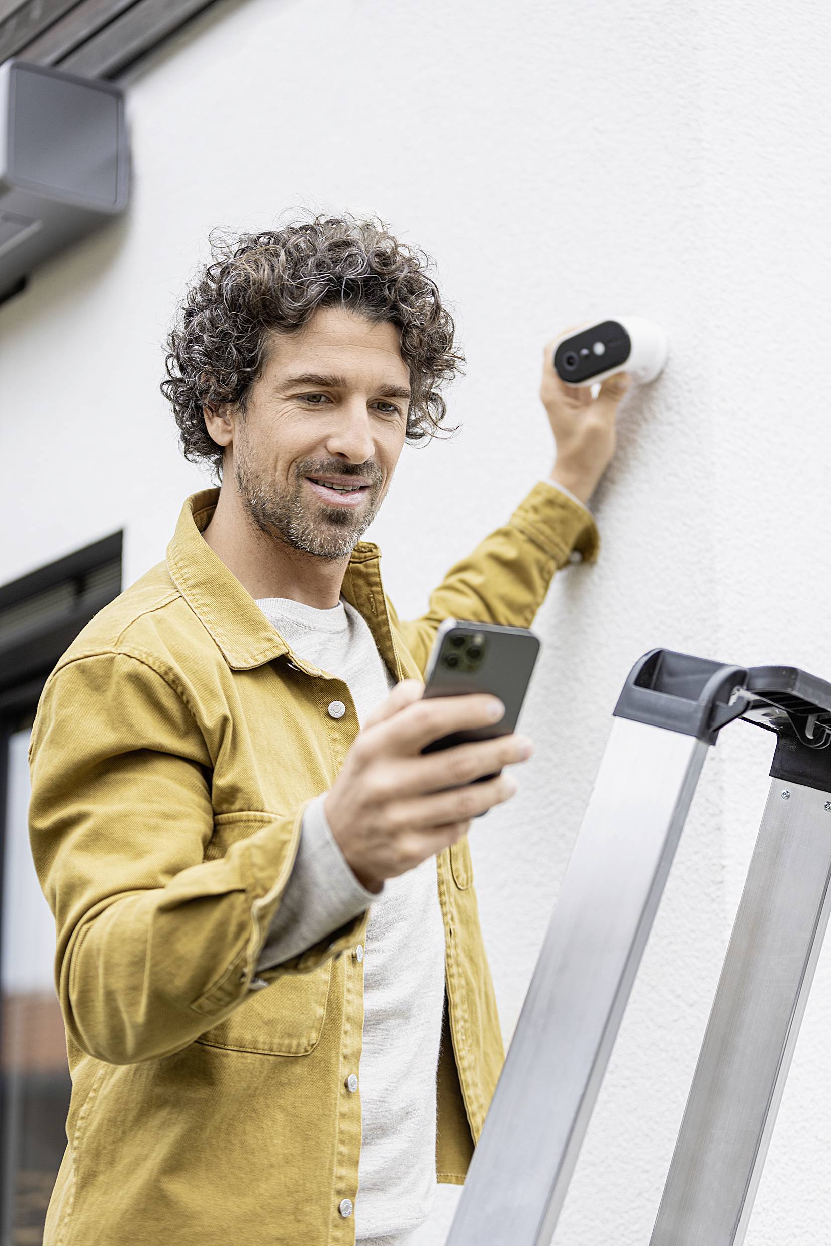 A man is attaching a security camera to a house wall while holding a smartphone. A ladder is standing next to him.