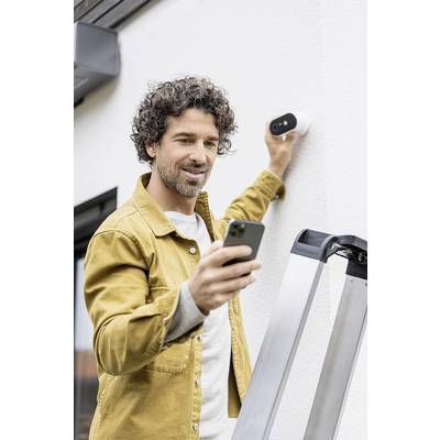 A man is attaching a security camera to a house wall while holding a smartphone. A ladder is standing next to him.