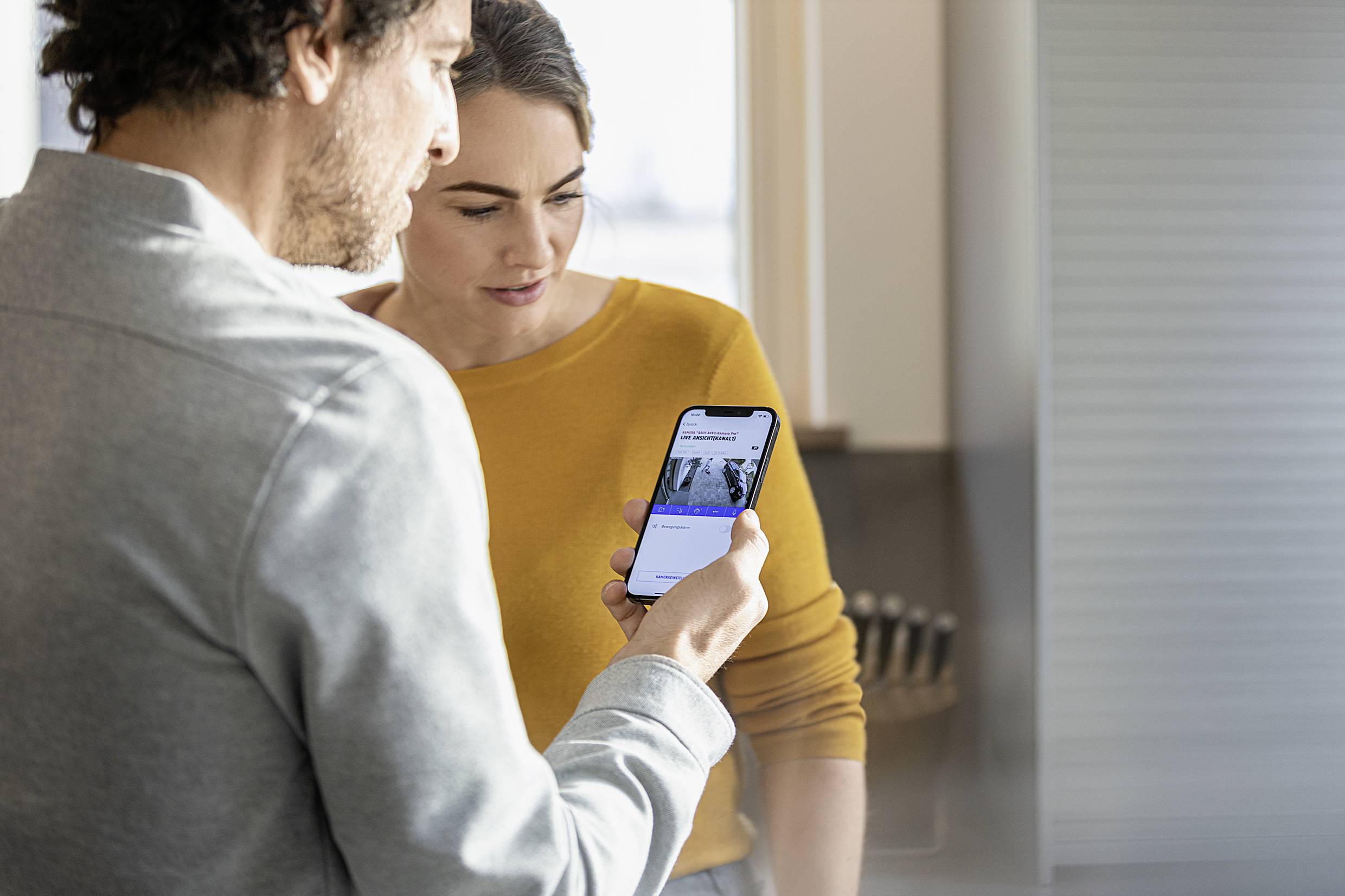 Two people are examining a smartphone. The woman is wearing a yellow jumper, and the man has on a grey top. They appear focused.