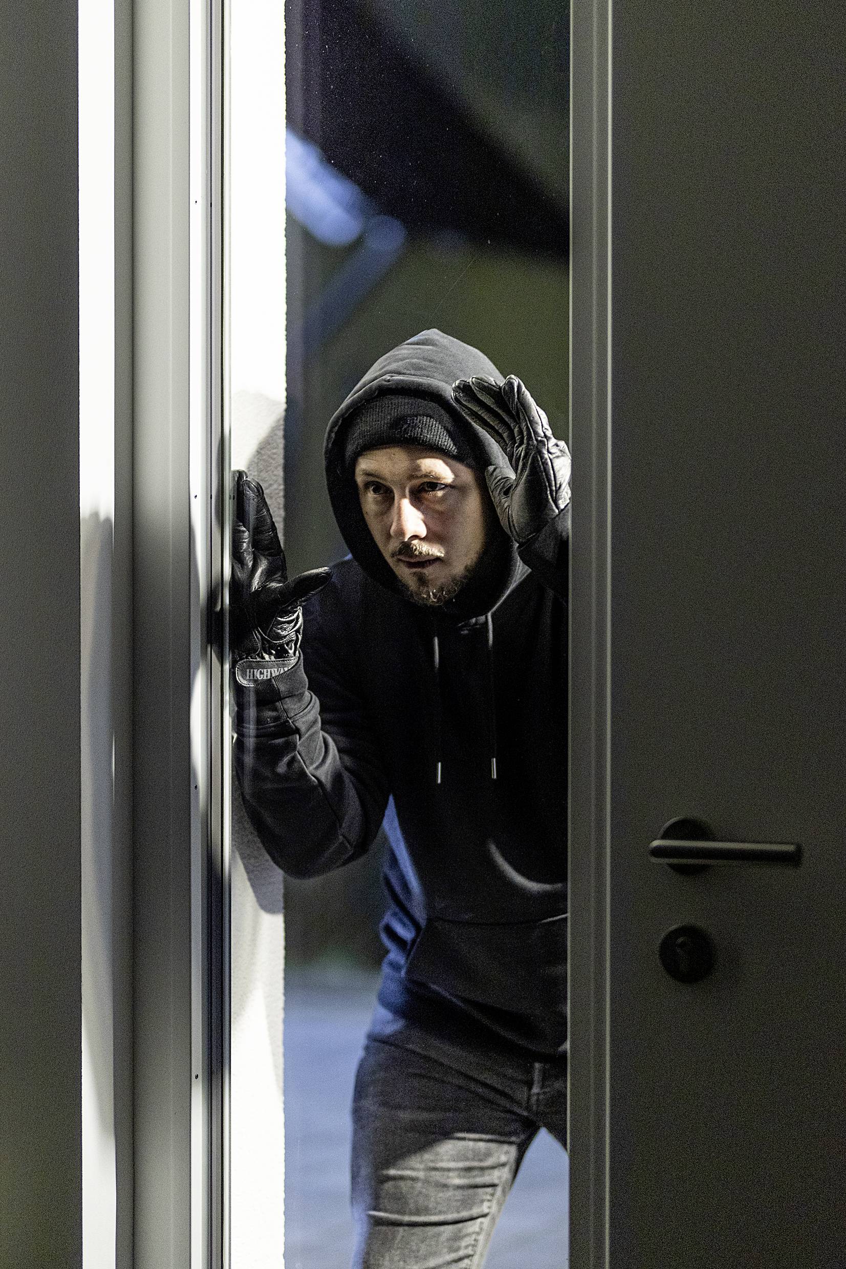 A man in dark clothing and a hood is peering through an open door, as if he intends to break into a building.