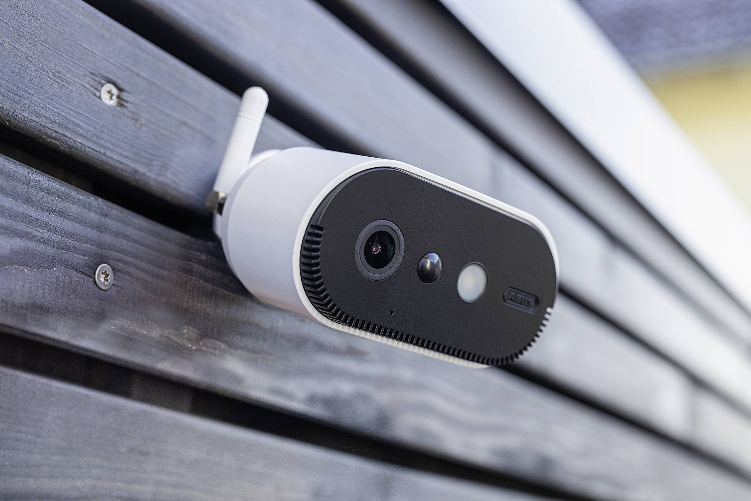 A surveillance camera is mounted on a wall. It has a white antenna and a black front panel with a lens and sensors.