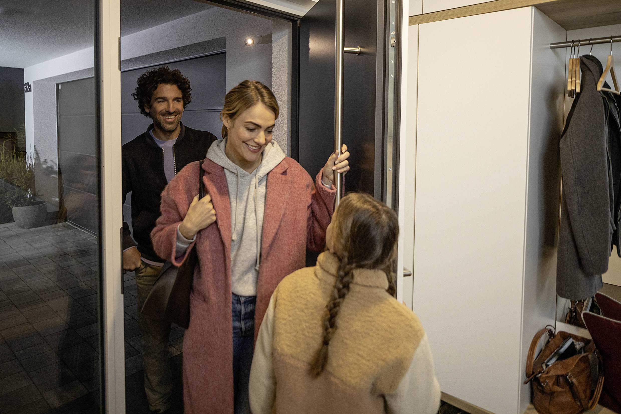 A woman in a pink coat enters a house with a smile, accompanied by a man in the background. A child greets her at the door.