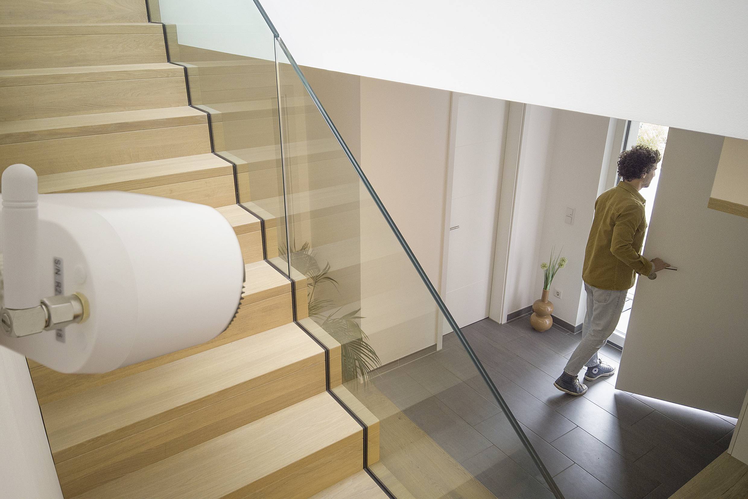 A person opens the front door, with a security device installed in the foreground. The staircase is made of wood with a glass banister.