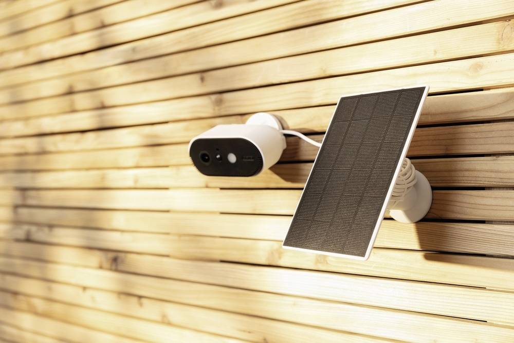 A surveillance camera with a solar panel is mounted on a light wooden lattice wall and illuminated by sunlight.