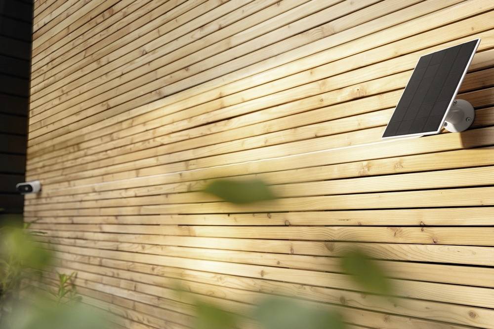 Solar panel mounted on a wooden facade, illuminated by sunlight. A camera is also attached to the wall.