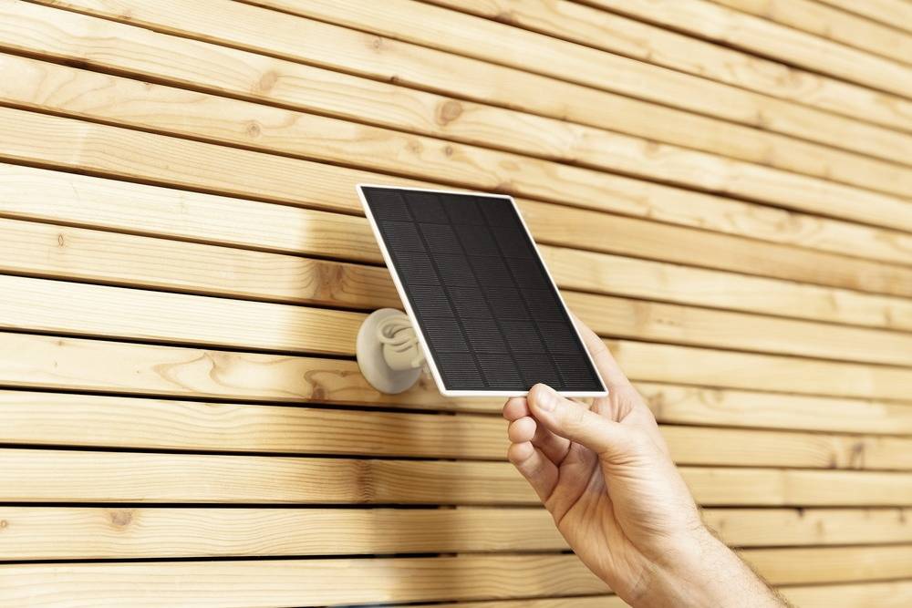 A hand is attaching a small solar panel to a light brown, vertically striped wooden surface.