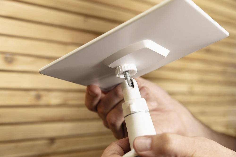 A person is attaching a rectangular, white solar panel to an adjustable mount in front of a wooden wall.