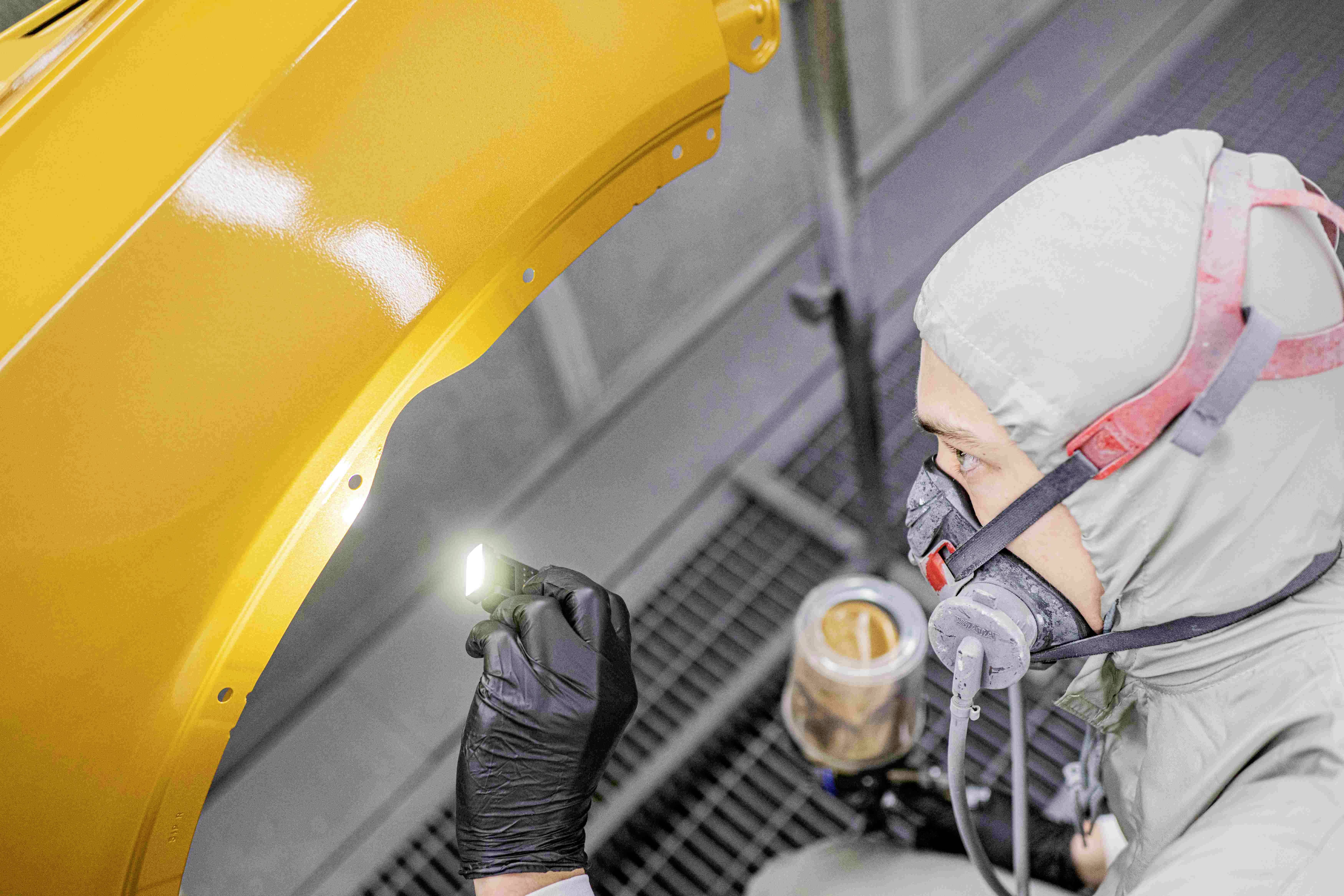 A person in protective clothing carefully inspects a yellow automotive component surface in a workshop environment, illuminated by a small lamp.