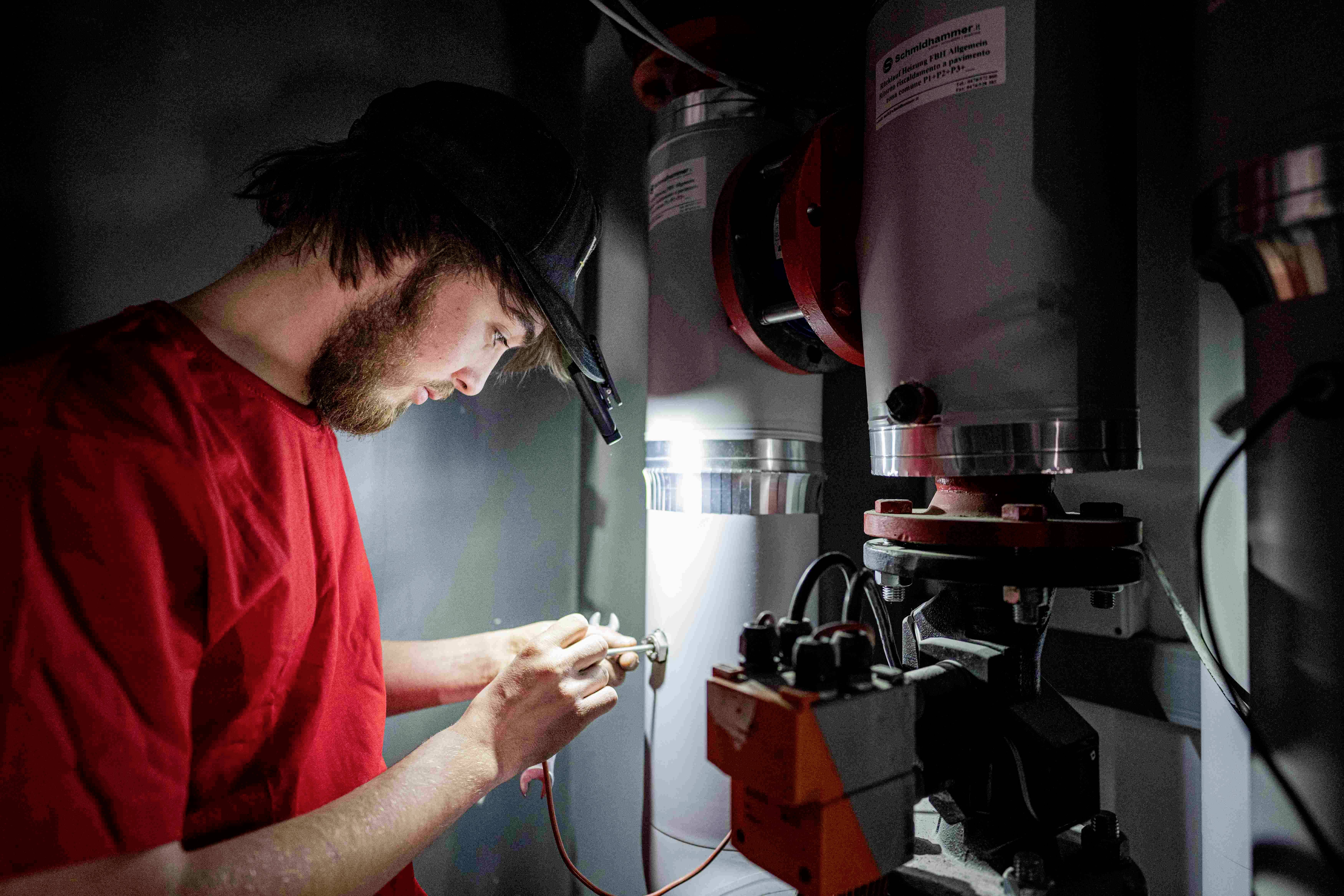 A man in workwear repairs technical equipment in a dimly lit room, focused on his task.