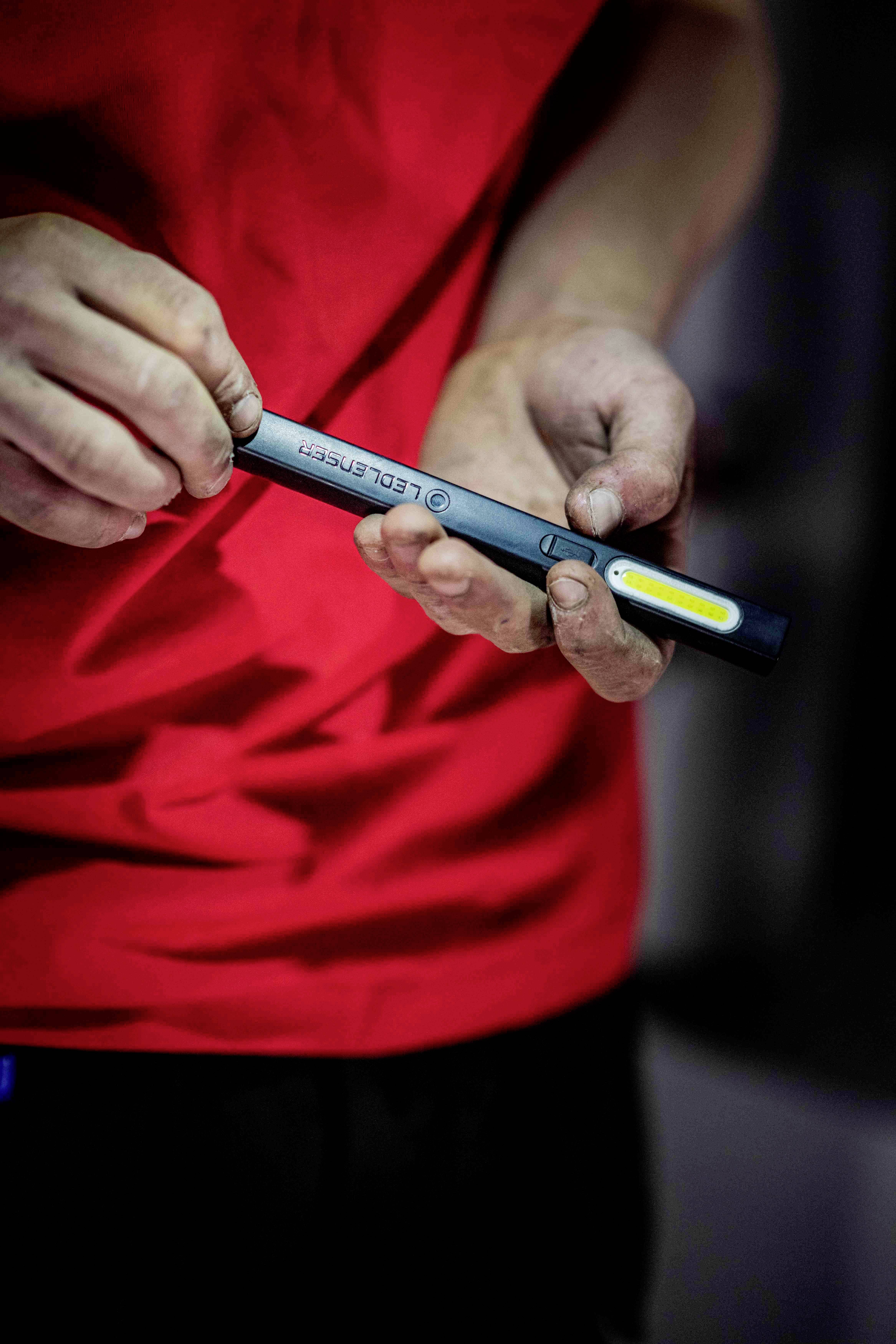 A person in a red T-shirt is holding an LED pen with torch functionality. Their hands are slightly dirty.