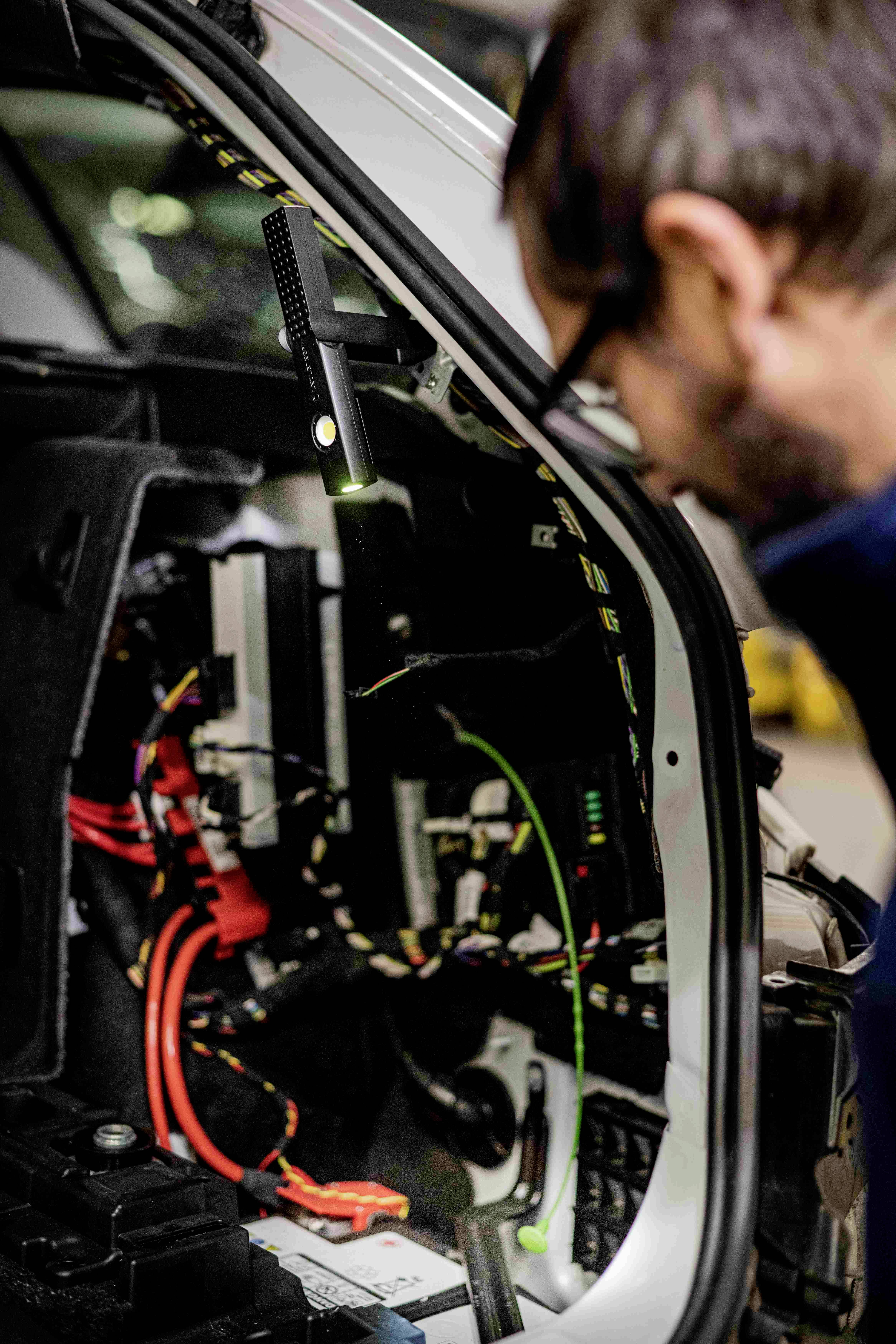 A man is inspecting the open boot of a car, with electrical wiring visible. A torch is shining on the components.