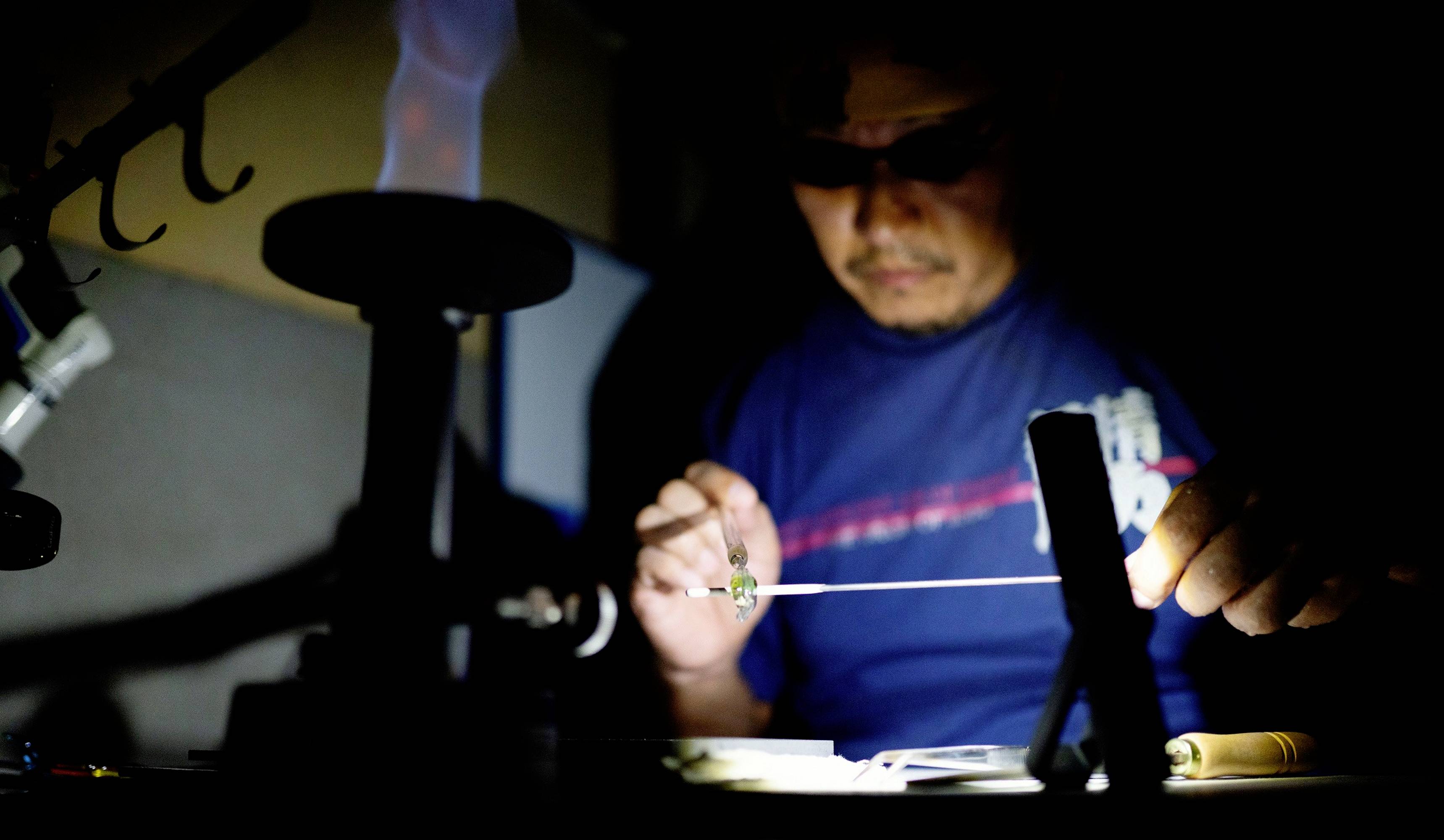 A man is working intently with a thin wire and flame in a dimly lit room, indicating a manual craft skill.