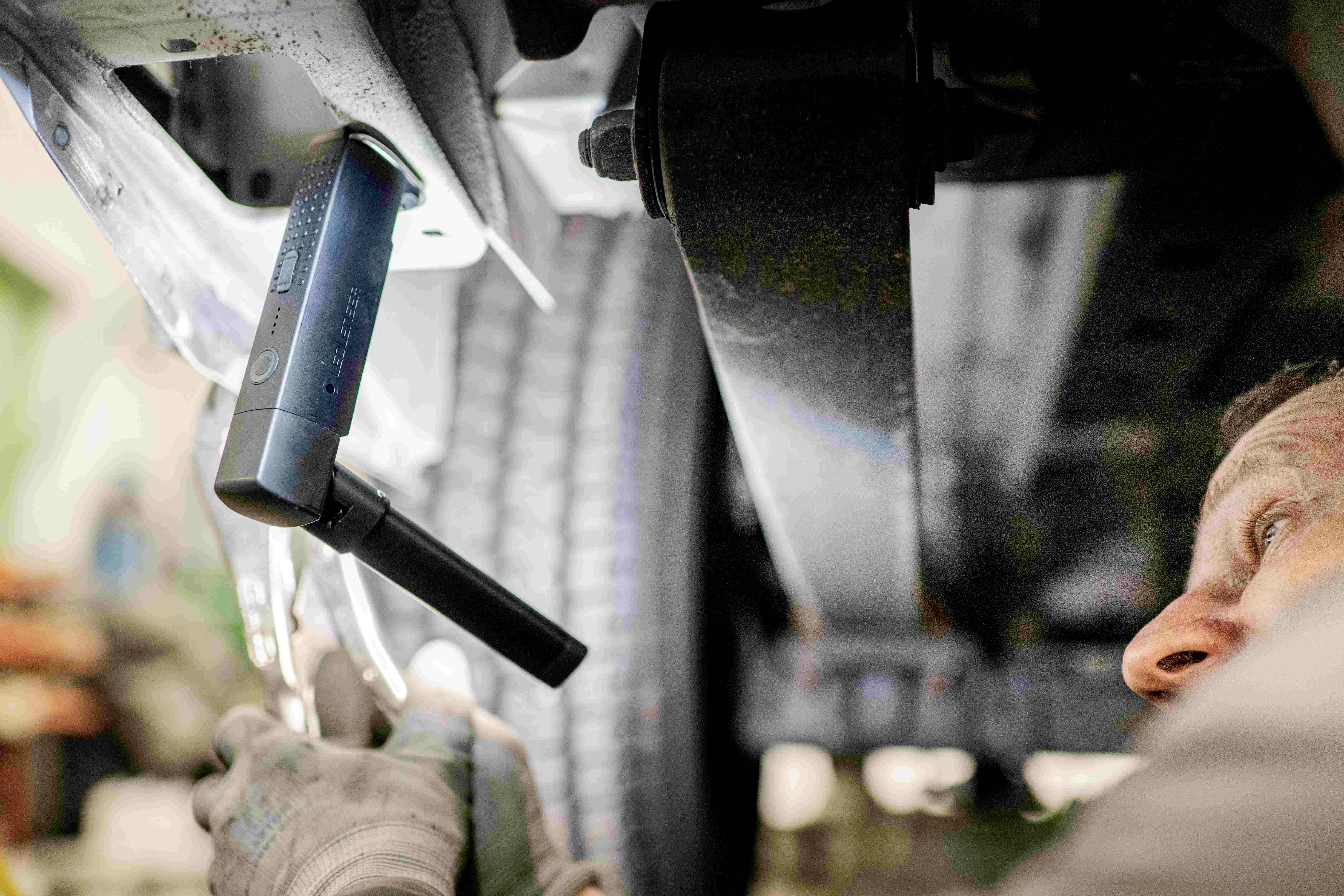 'A mechanic repairs a vehicle from underneath using a torch and tools.'<br><br>Note: In British English, 'torch' is used instead of the American 'flashlight', and the phrasing is slightly adjusted to sound more natural.