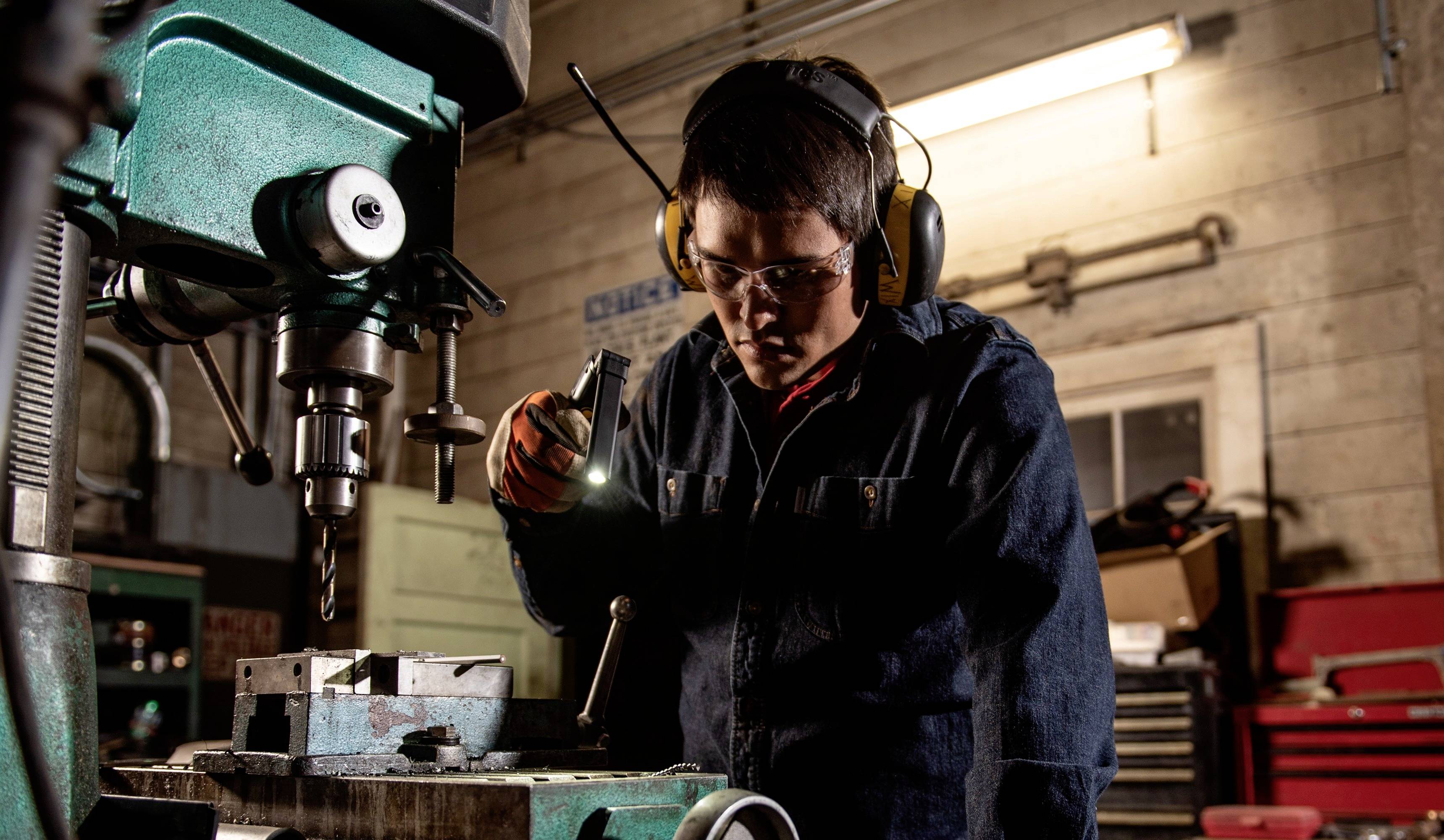 A person is working on a milling machine in a workshop. They are wearing safety glasses and ear protection.