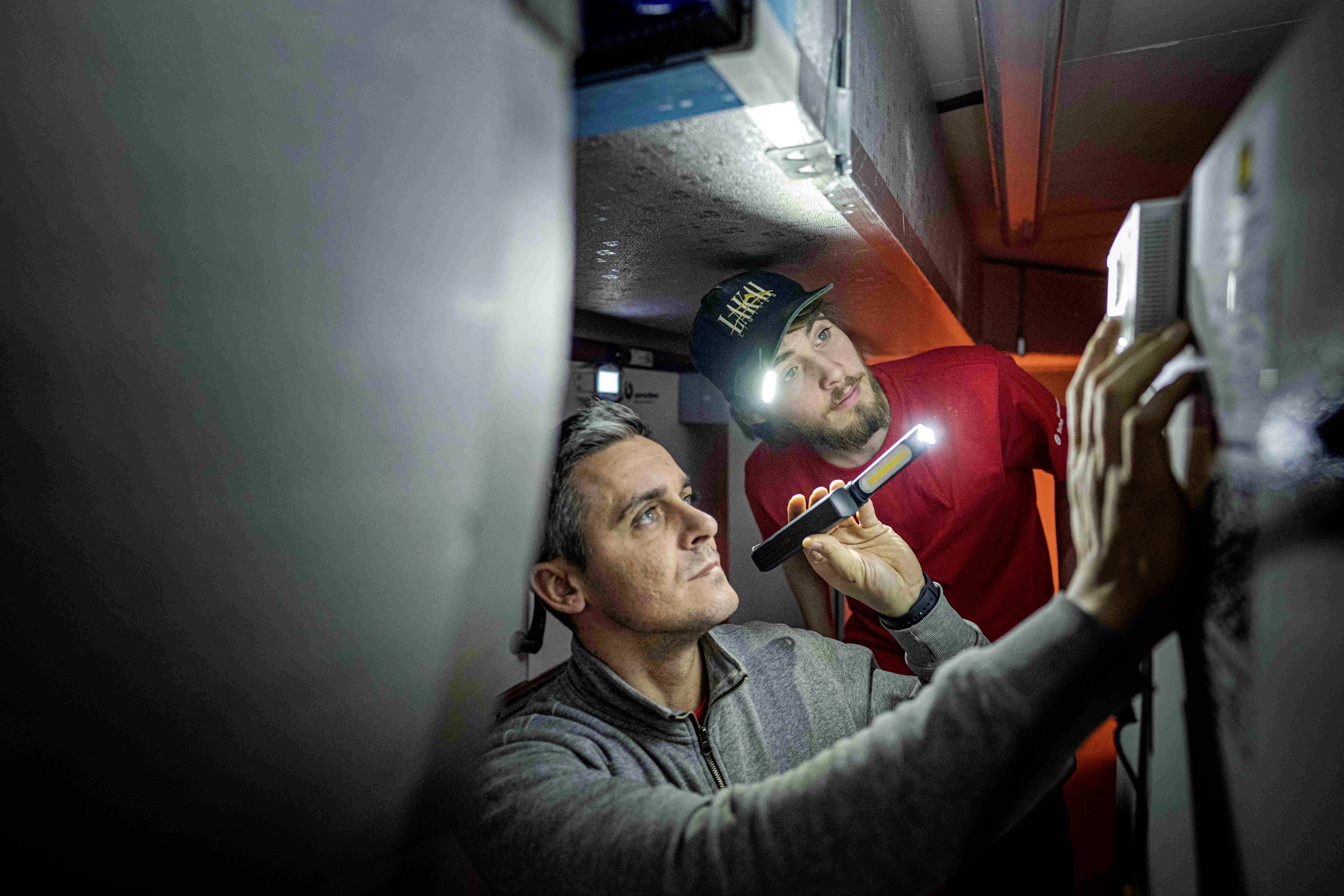 Two men are working in a confined space; one is holding a torch, the other is examining an electrical switchboard.