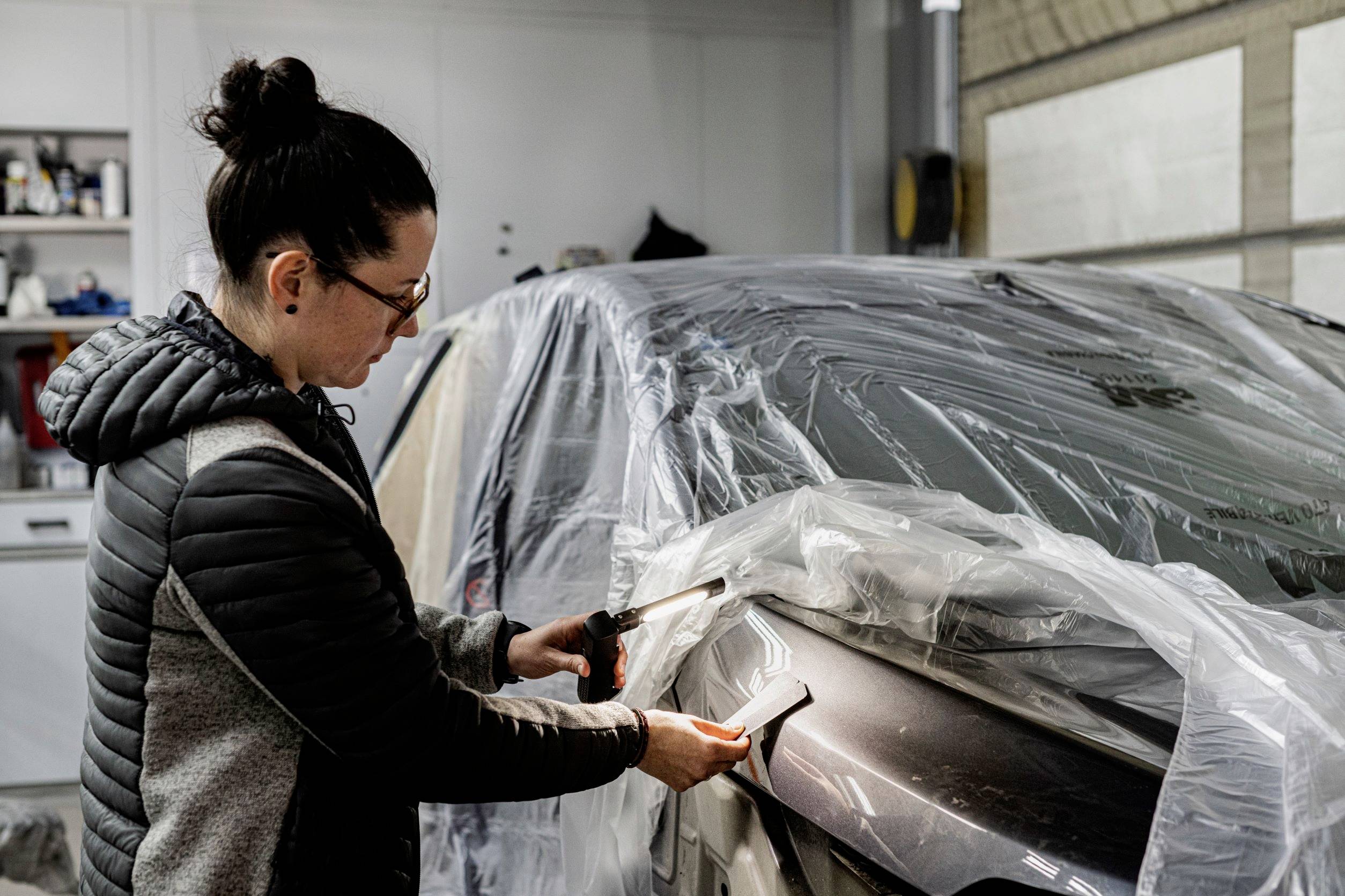 A woman wearing glasses is repairing a car in a garage; she is using plastic material and a tool on the bumper.