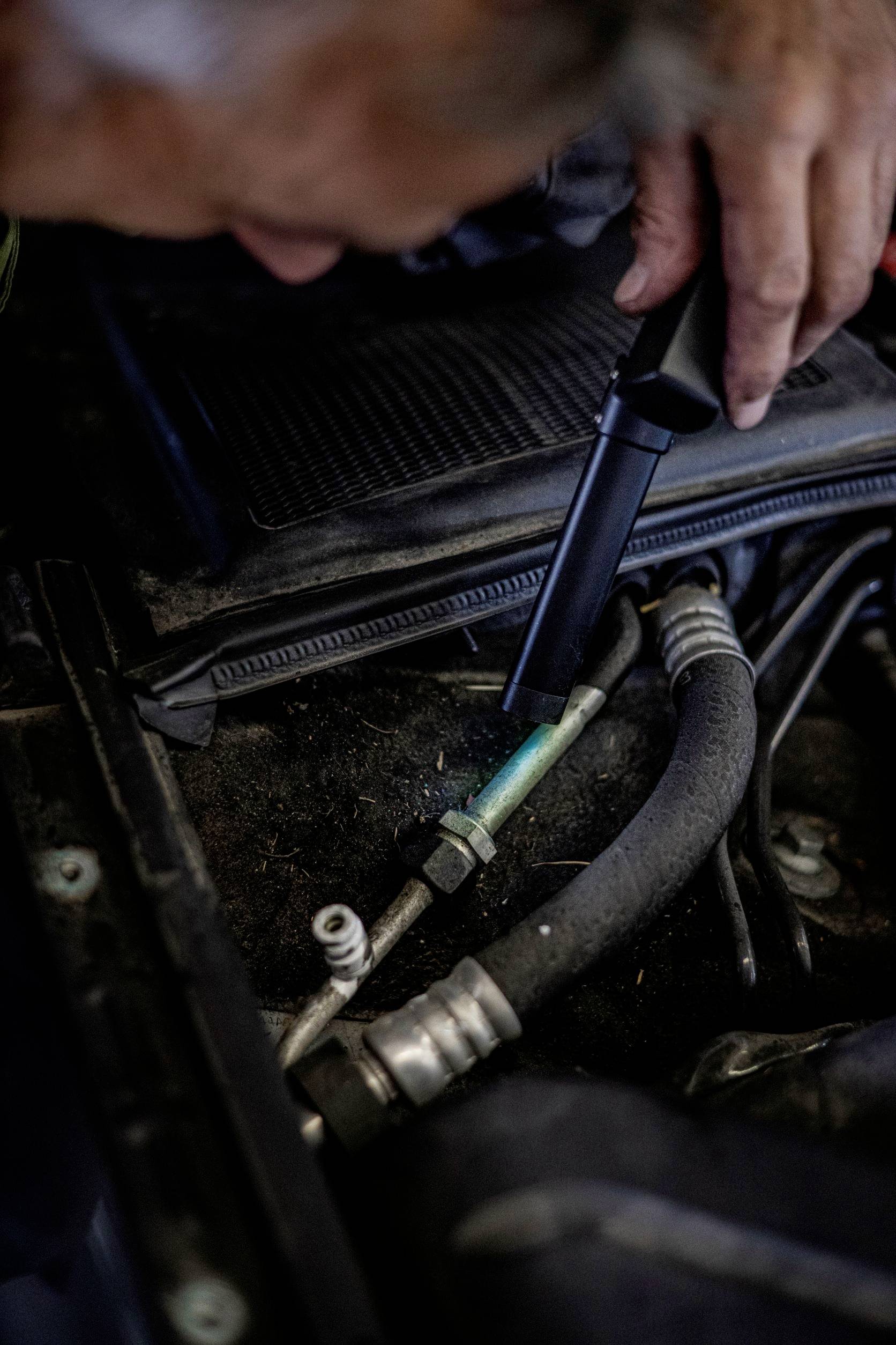A mechanic inspects a car part in the engine bay with a torch. Focus on hoses and connections.