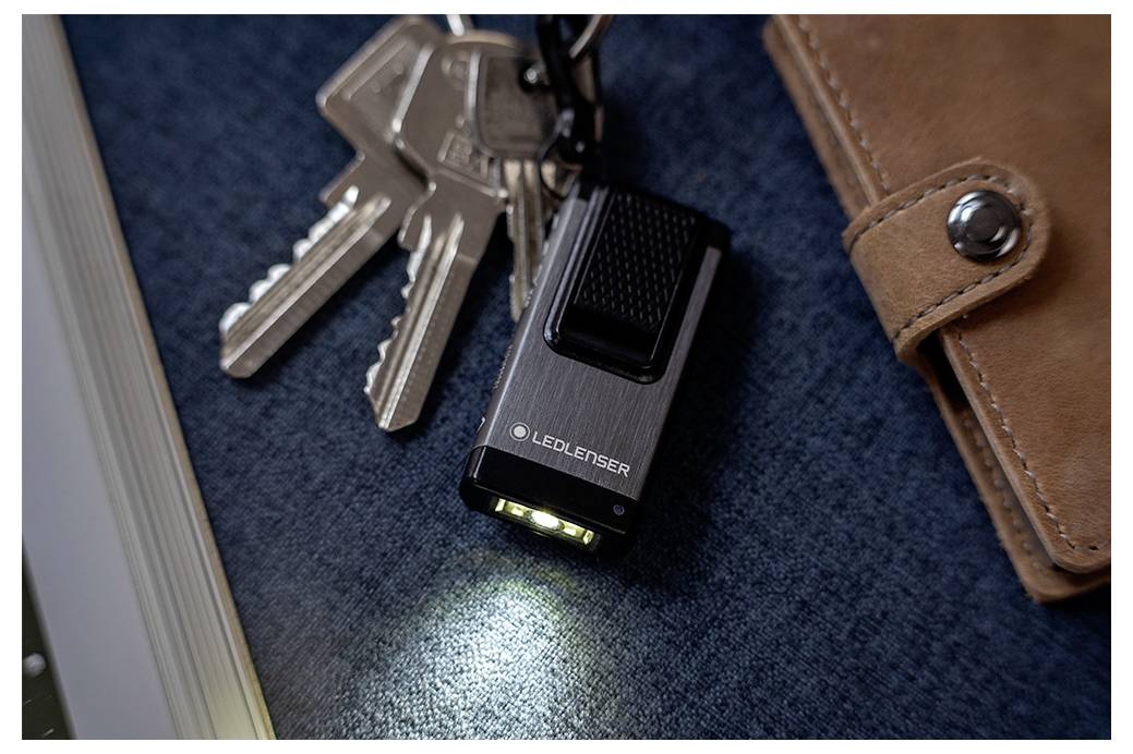 Small LEDLENSER flashlight shining on a blue surface beside keys and a brown leather wallet.
