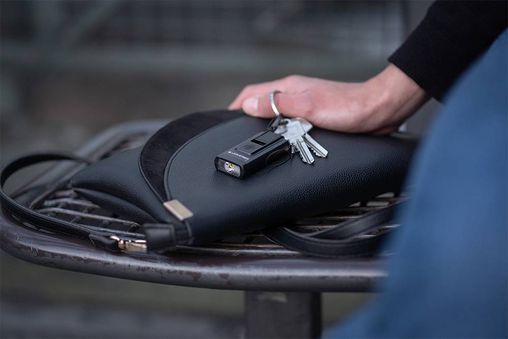 A hand is holding a bunch of keys with a torch on a black bag resting on a bench.