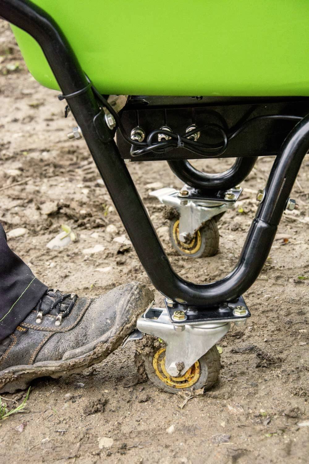 'A worker in boots is manoeuvring a wheelbarrow across a muddy, uneven ground. The wheels of the barrow are partially sunk into the mud.'
