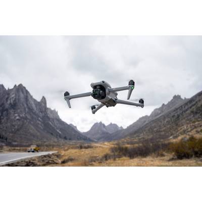 A drone is flying over a mountainous landscape with a cloudy sky. In the background, rugged peaks and a car on a road are visible.